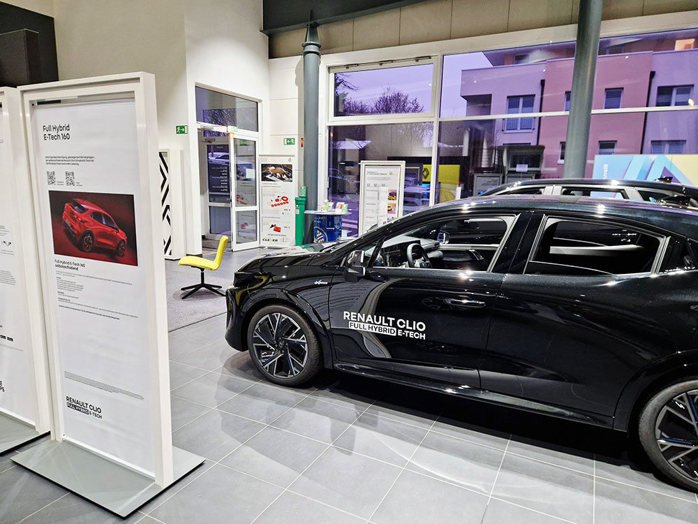 Automobile Clever Service Schwarzer Renault Clio E-Tech Full Hybrid in einem Autohaus, daneben ein Informationsaufsteller und ein gelber Stuhl.
