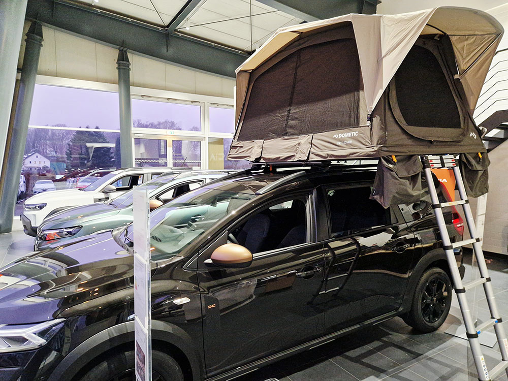 Automobile Clever Verkauf Schwarzes Dacia-Fahrzeug mit aufgeklapptem Dachzelt und Leiter im Autohaus, im Hintergrund weitere ausgestellte Fahrzeuge.