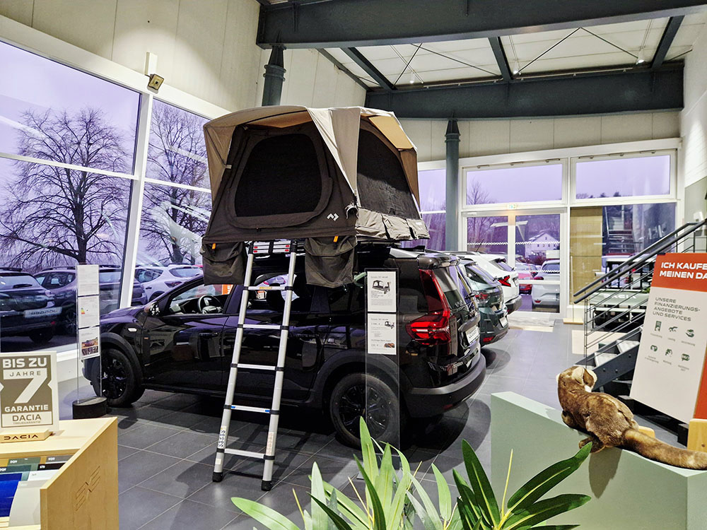 Automobile Clever Verkauf Dacia-Fahrzeug mit aufgebautem Dachzelt im Autohaus Clever, umgeben von weiteren ausgestellten Fahrzeugen und Informationen zum Zubehör.