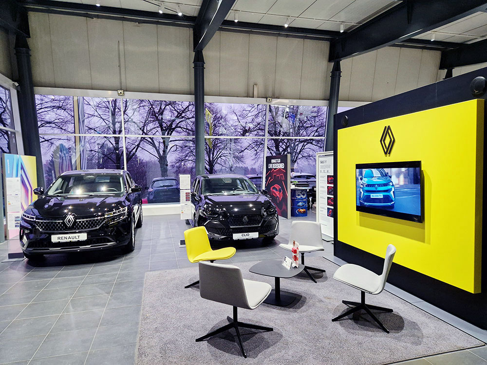 Automobile Clever Werkstatt Showroom vom Autohaus Clever mit der Marke Renault mit zwei ausgestellten Fahrzeugen, Sitzbereich und großem Renault-Logo an einer gelben Wand.