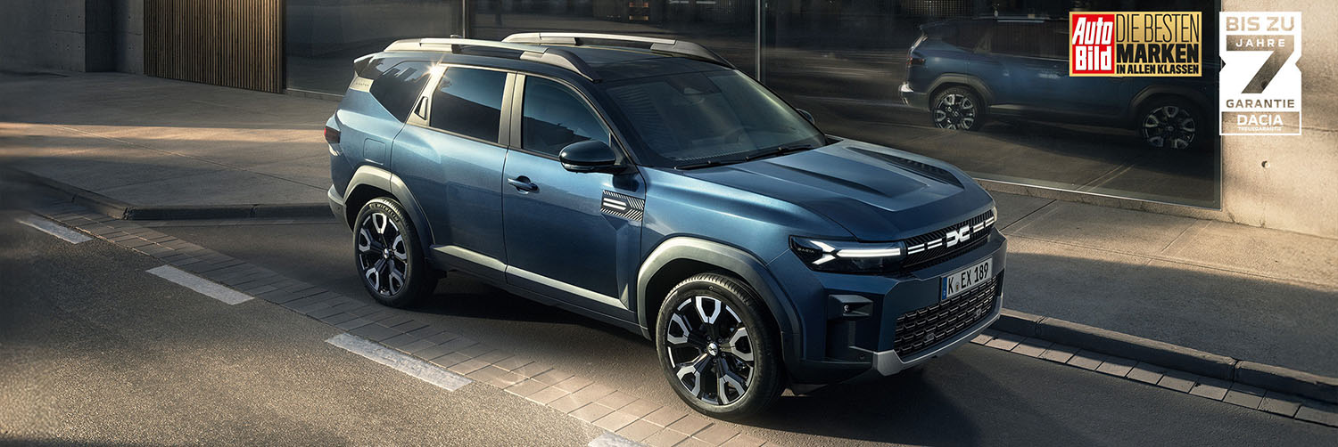 Ein blauer Dacia Bigster steht in der Sonne vor einem modernen Gebäude. Das SUV ist aus der Front-Seiten-Perspektive zu sehen und zeichnet sich durch markante LED-Scheinwerfer, schwarze Radlaufverkleidungen und sportliche Leichtmetallfelgen aus. Im Hintergrund steht ein weiteres Fahrzeug. Im oberen rechten Bildbereich sind Hinweise auf Auszeichnungen und die Dacia-Garantie zu sehen.